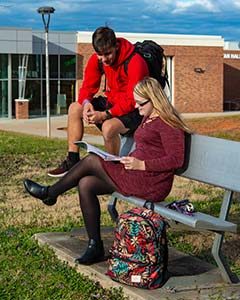 Two WPCC students sit on a bench on the campus grassy commons, talking and studying