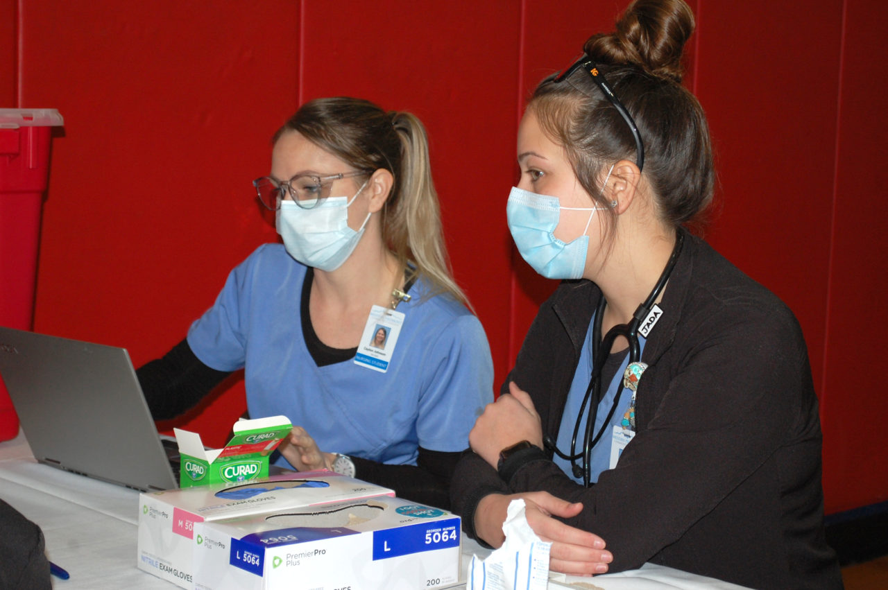 Two nursing students in scrubs and masks look at a laptop at the clinic site.