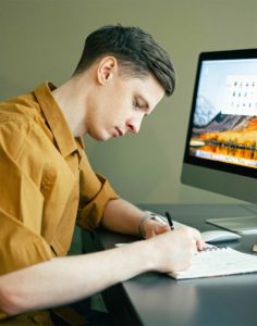 A male student takes notes while sitting at a computer