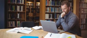 Photo of a man working at a computer with several college textbooks open around him