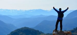 Image of a male standing on a rock with both fists in the air looking at the mountains.