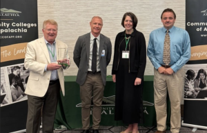 4 people smiling for a photo with one person holding an award and with a banner on each side.